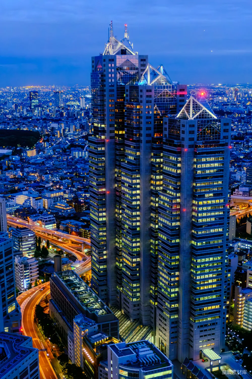 这张图片展示了夜晚的城市景象，中央是一栋高耸的摩天大楼，灯火辉煌，周围是灯火通明的街道和建筑。背景是远处的建筑群，天空呈现出深蓝色。整体画面充满了现代都市的繁华与活力。