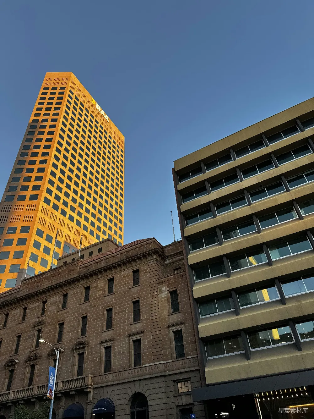 这张图片展示了城市中现代与历史建筑的对比，高耸的现代玻璃幕墙建筑在夕阳的映照下呈现出金黄色，而下方的建筑则保留了经典的历史风貌。天空的蓝色与建筑物的色彩形成鲜明对比，营造出一种独特的城市景象。适合用于城市规划、建筑设计或城市风光的主题。