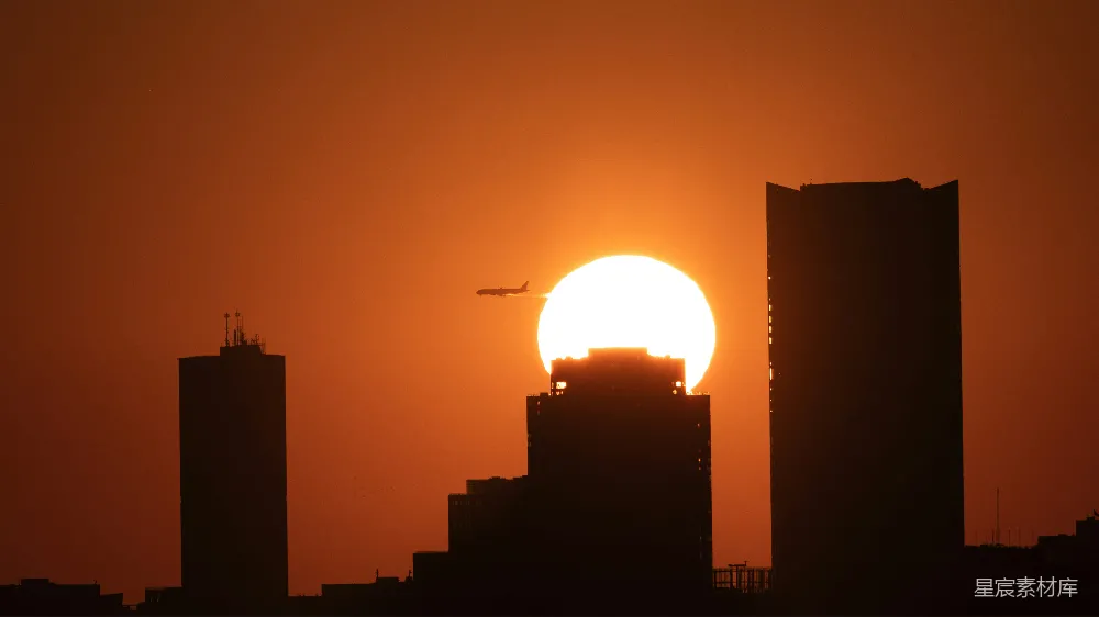 这张图片捕捉到了城市天际线的日落时刻，夕阳的光辉从高楼大厦之间洒下，一架飞机在空中飞行，留下一道剪影，整个场景充满了宁静与动态的结合。