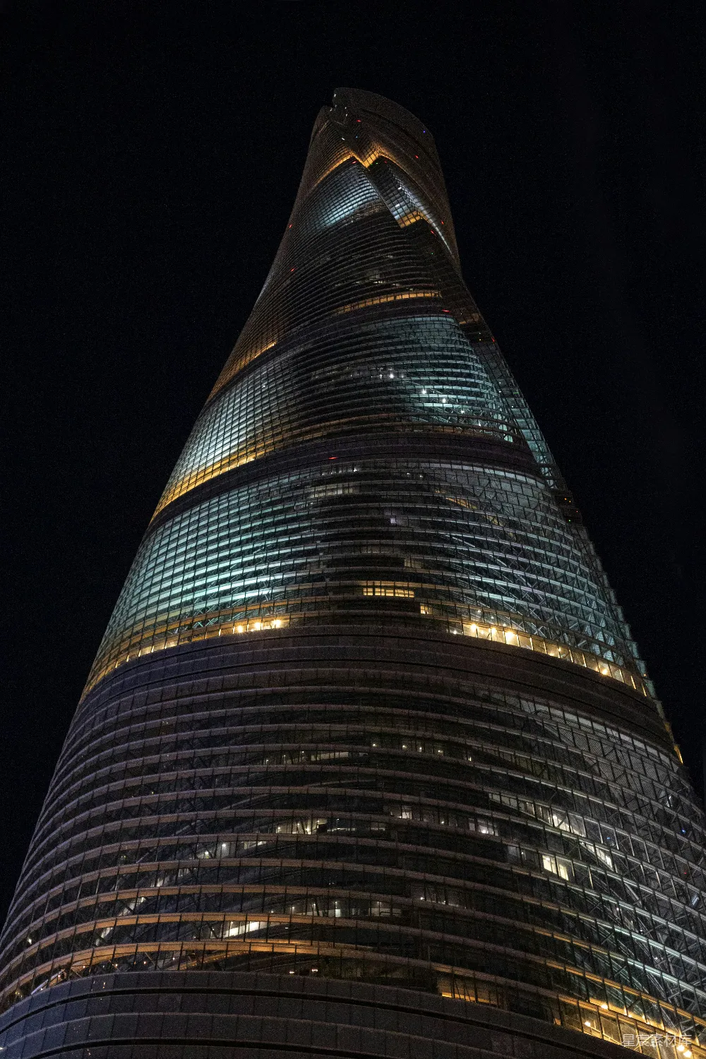 这张图片展示了上海中心大厦在夜晚的壮观景象。建筑的玻璃幕墙在灯光的映衬下显得晶莹剔透，曲线设计在黑暗中尤为突出，展现出都市的现代感和科技感。这幅图像非常适合用作城市风光、建筑艺术或都市生活的展示。