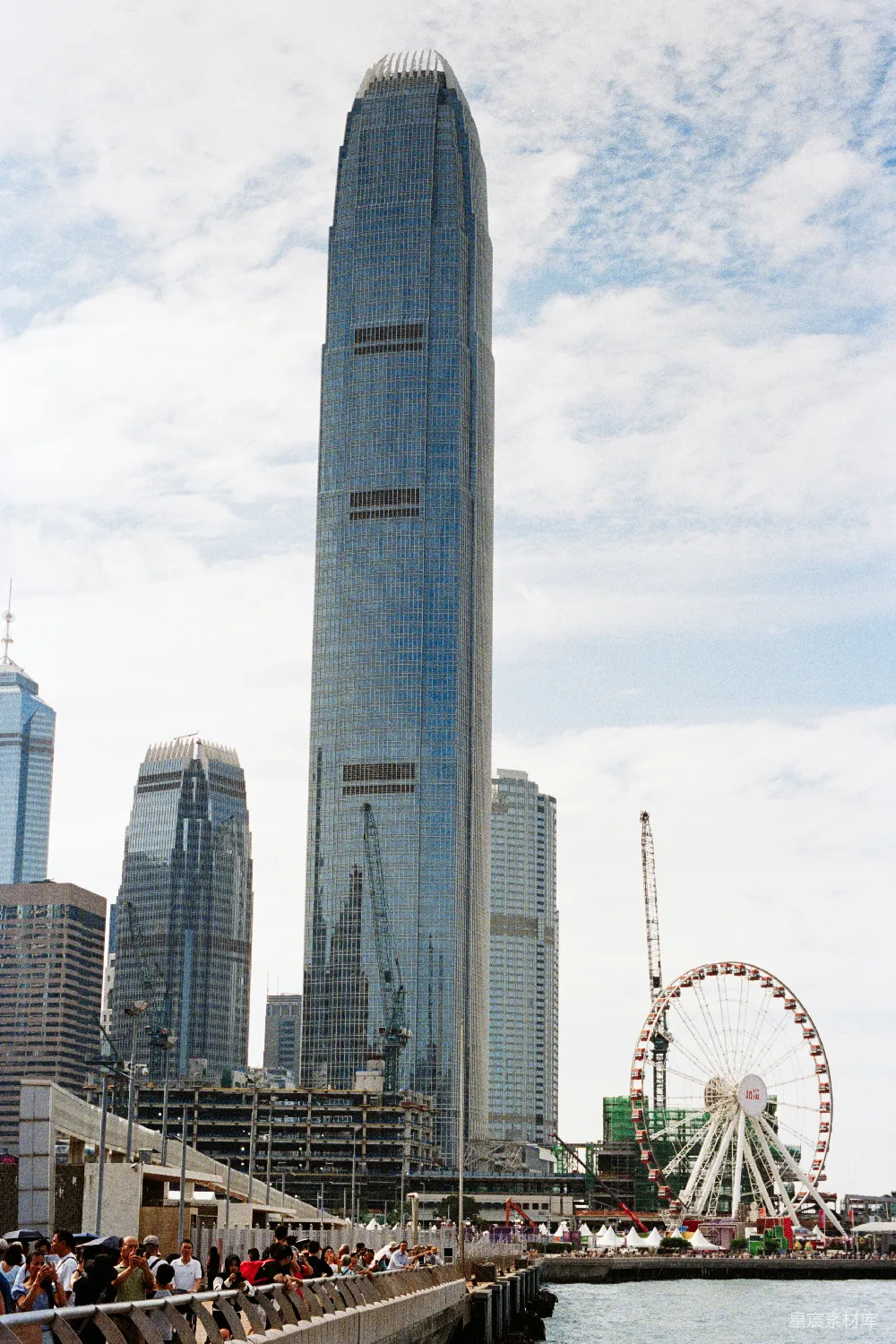 这张图片展示了香港著名的天际线，背景中可以看到一座高耸的现代化摩天大楼和一个大型摩天轮。前景中，游客们聚集在观景台，享受着城市的美景和壮观的建筑景色。天空中散布着几朵白云，为整个场景增添了宁静的氛围。这张照片适合用于城市旅游、现代建筑和观光