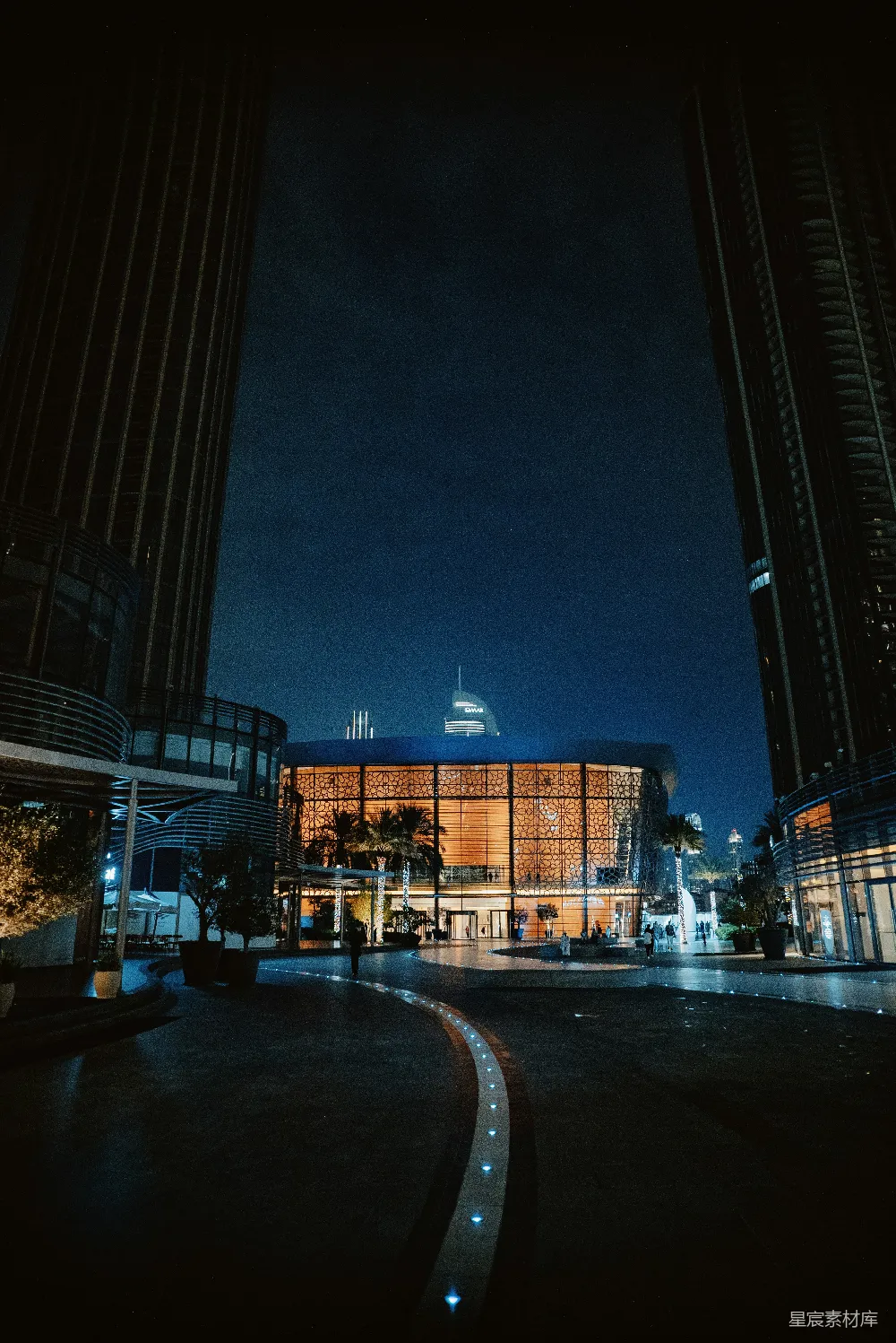 这张图片展示了一座城市的夜景，画面中央是一座被灯光照亮的建筑，周围环绕着现代风格的高楼。地面上的灯光线条形成了引导线，增加了画面的深度和动感。远处隐约可见的城市天际线和高耸的建筑，营造出一种繁华而宁静的氛围。