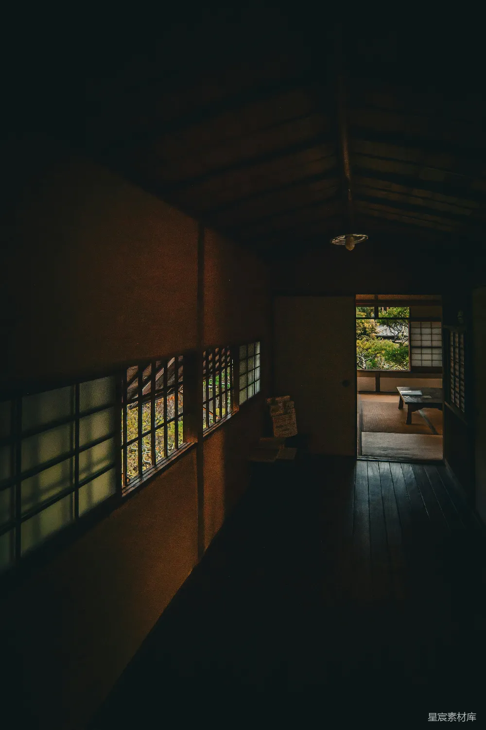 这张图片展示了一个日式的房间，室内光线昏暗，只有透过窗户和门的自然光透进来。墙壁上是传统的日式障子门，地板是木质的，整体色调偏暗，营造出一种宁静而神秘的氛围。这种场景适合用于描述安静的阅读角落、冥想空间或者展示传统日式建筑的室内设计。