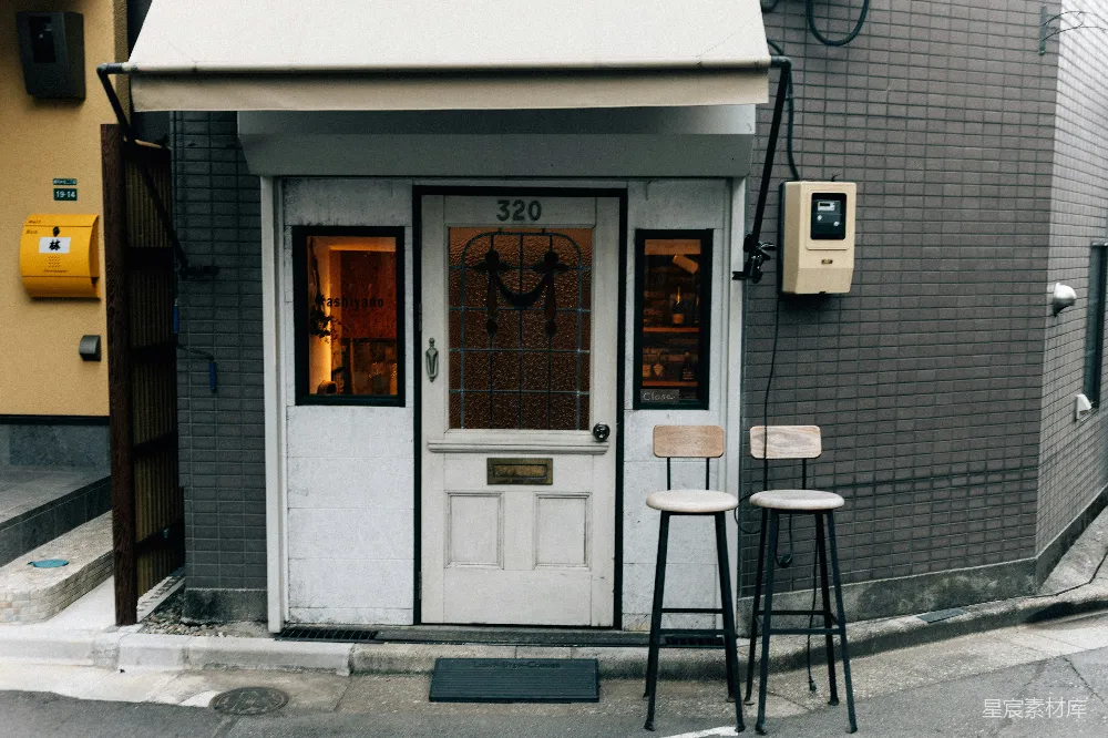 这张图片展示了一家位于街道旁的复古风格咖啡店门廊。门廊前摆放着两把高脚凳，透过玻璃门可以看到店内的温馨灯光和装饰。门廊的设计简洁而具有特色，适合用来拍摄店铺的街景照片，或作为城市生活场景的一部分。