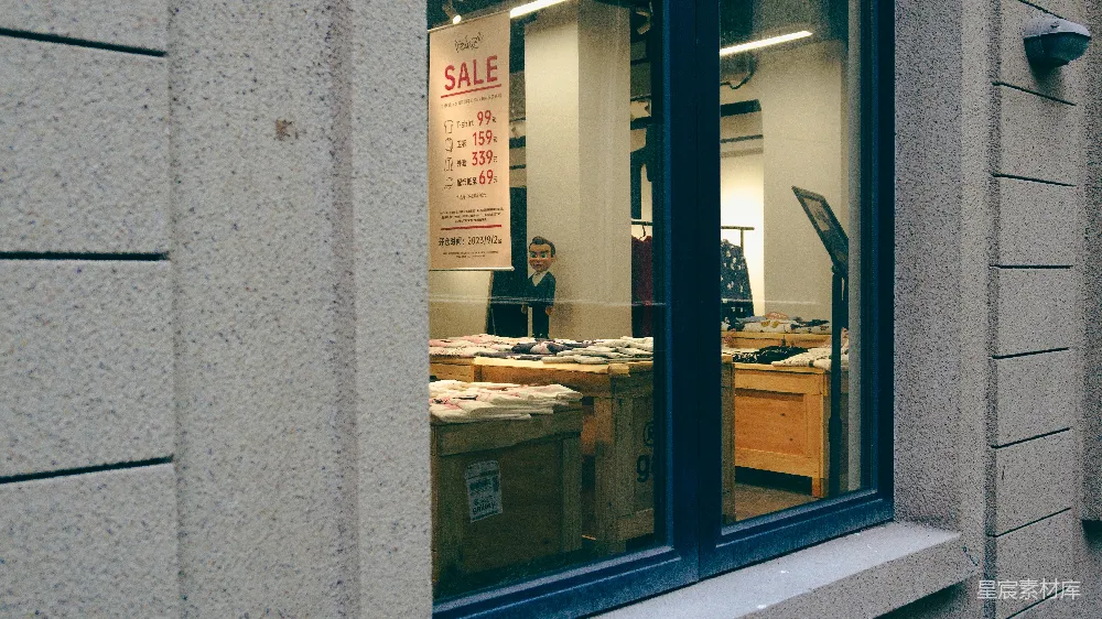这张图片展示了一家商店的橱窗，橱窗内有一个促销的海报，显示了打折的商品。橱窗外是灰色的墙壁，窗户的玻璃反射出室内的情况。海报上方醒目地写着“SALE”，并列出了各种商品的原价和打折后的价格。这种场景常见于商店促销期间，吸引顾客进店购买。