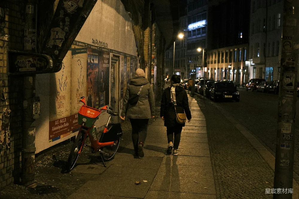 这张图片展示了一个夜晚的城市街景，两名行人沿着人行道行走，背景是昏暗的街道和高楼。墙上有各种海报和涂鸦，左侧停着一辆橙色的自行车，街道两旁有路灯照亮，营造出一种宁静而神秘的氛围。适合用于城市生活、夜景和人文主题的素材。