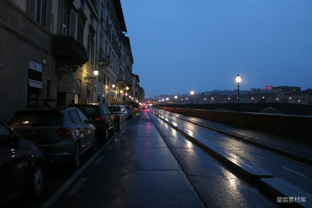 这张图片展示了一个宁静的雨夜场景，街道两旁停满了汽车，路灯和远处的建筑物投射出温暖的光芒，反射在湿润的地面上，营造出一种宁静而美丽的氛围。适合用于城市夜景、雨天主题或宁静氛围的设计素材。