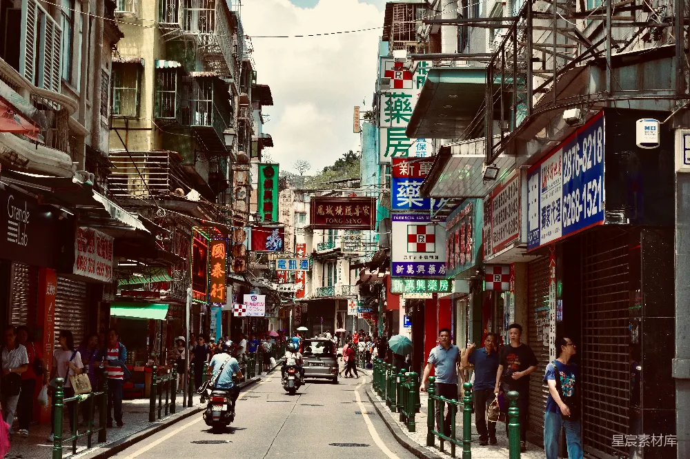 图片展示了一个亚洲城市的繁华街道，两旁是错落有致的商店和住宅楼，招牌色彩鲜艳，街道上人来人往，有行人、电动车和摩托车穿梭其间。阳光明媚，天空晴朗，营造出一种热闹的氛围。适合用于城市生活、亚洲风情或商业街道的宣传背景。