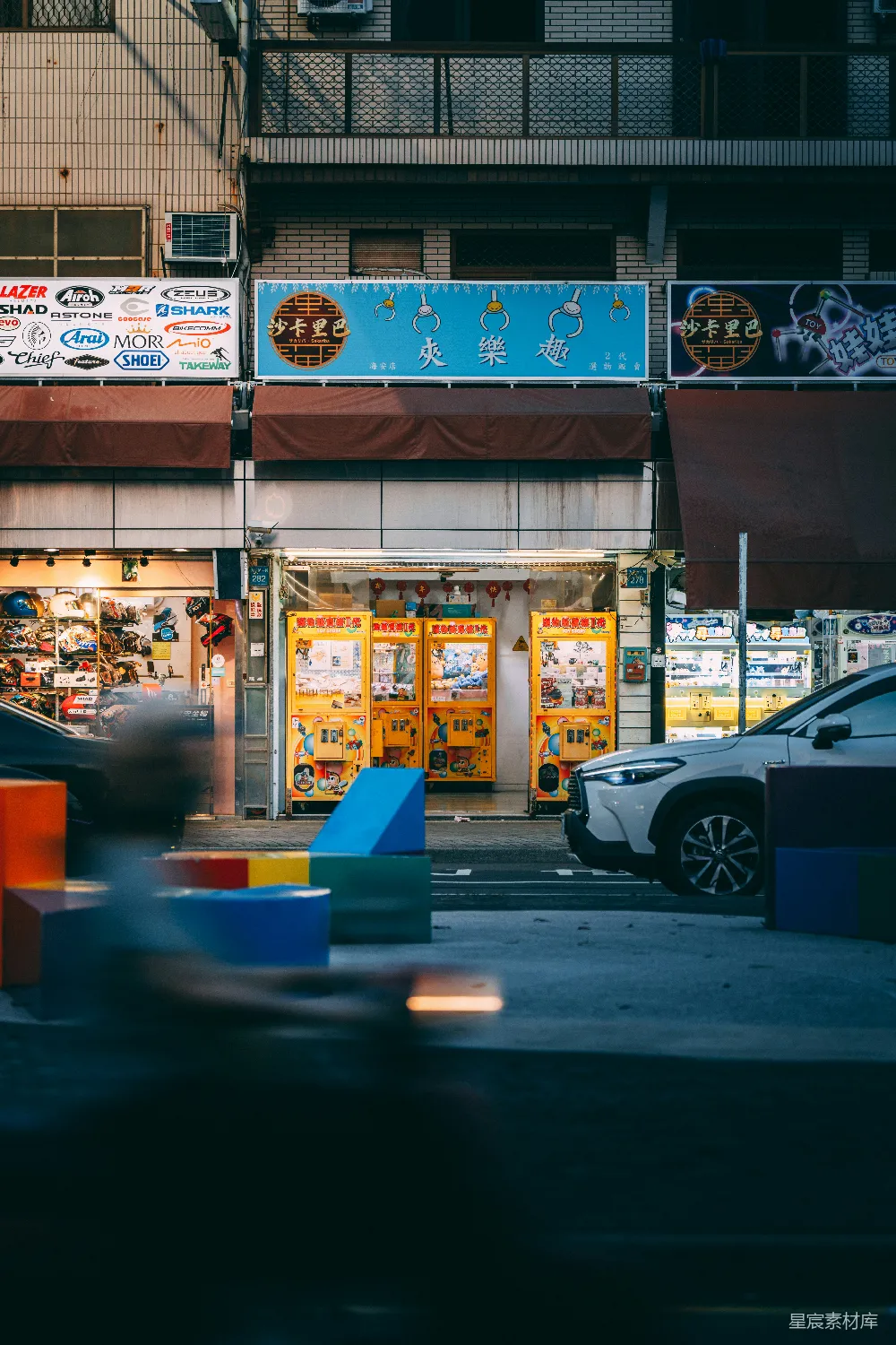 这张图片展示了一家位于城市街景中的夹娃娃机店。夜晚的灯光下，夹娃娃机的黄色灯光显得格外醒目，背景是建筑物和招牌，前景中有一些彩色的交通锥。整个场景充满了都市的夜晚氛围，适合用于休闲娱乐或城市生活主题的素材。