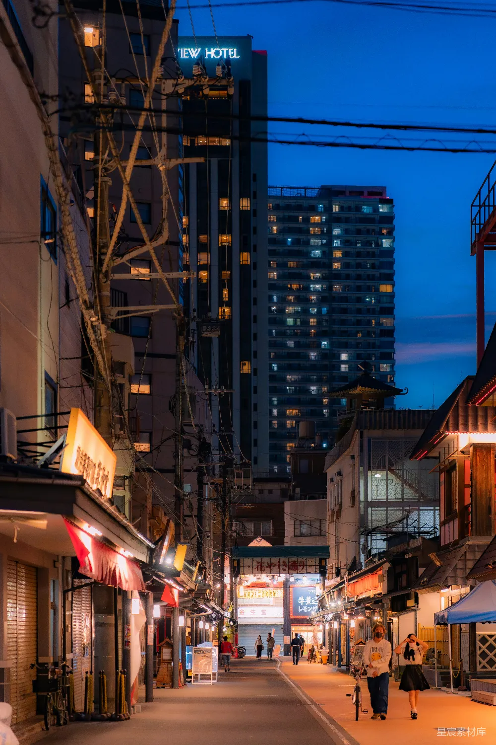 这张照片展示了夜晚城市的街景，街道两旁是亮着灯光的商店和住宅楼。霓虹灯与街灯交织出一幅迷人的夜景图，远处的高楼和近处的低矮建筑形成对比。街道上行人和骑自行车的人点缀其间，显得生活气息浓厚。整体色调偏蓝，营造出宁静而充满活力的氛围。