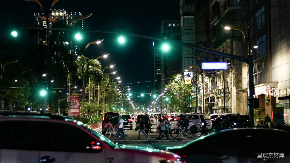这张图片展示了夜晚城市的一角，街道两旁的建筑物灯火通明，道路上车辆和行人交织，绿灯亮起，照亮了整个街道。路旁的棕榈树在灯光下显得格外醒目，营造出一种繁忙而充满活力的都市夜景氛围。