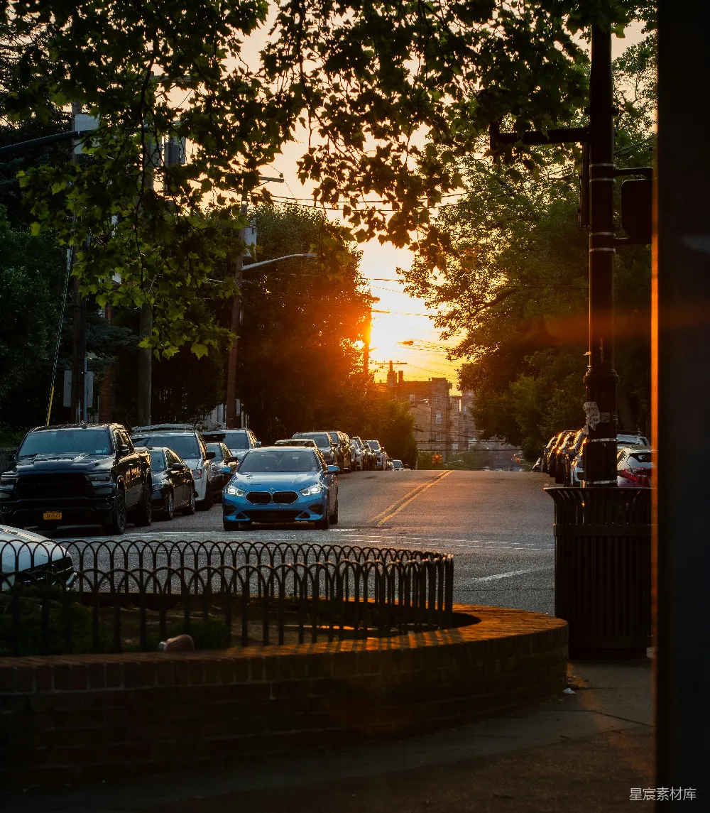 图片展示了一条宁静的街道，夕阳的余晖洒在道路上，树木的剪影与城市的轮廓相得益彰。街道两旁停满了车辆，远处的建筑在温暖的阳光下显得格外宁静。这幅画面给人一种平和、舒适的感觉，适合用于城市生活、自然风光或宁静时光的场景。
