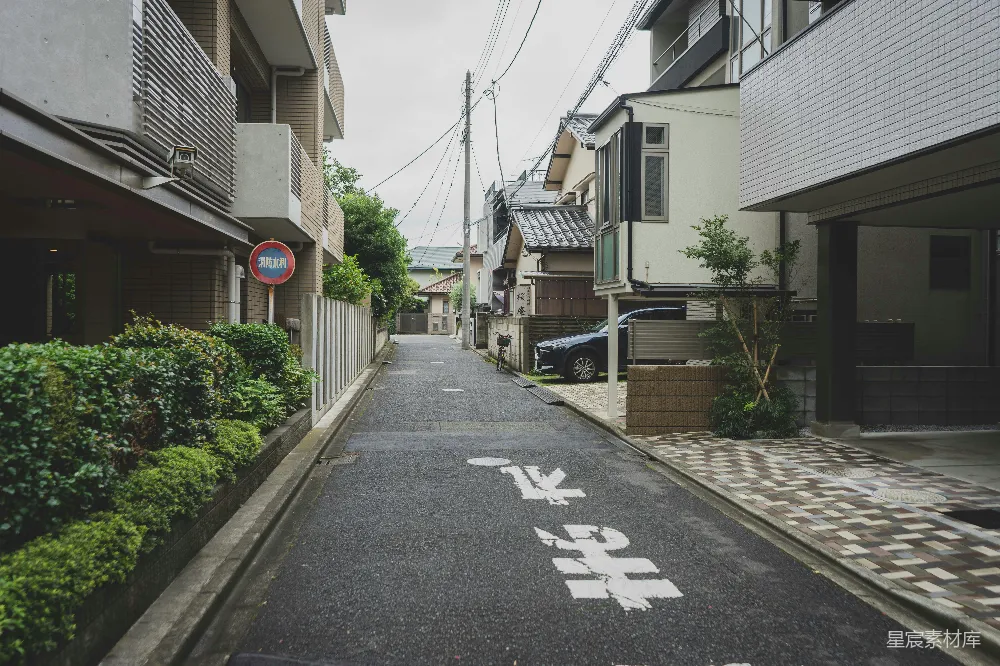 这张图片展示了一条安静的日本住宅区街道，两旁是现代化的住宅建筑，路面干净整洁，路标清晰，两边有整齐的绿植。整体氛围宁静，适合用于描述都市生活中的安静居住环境。