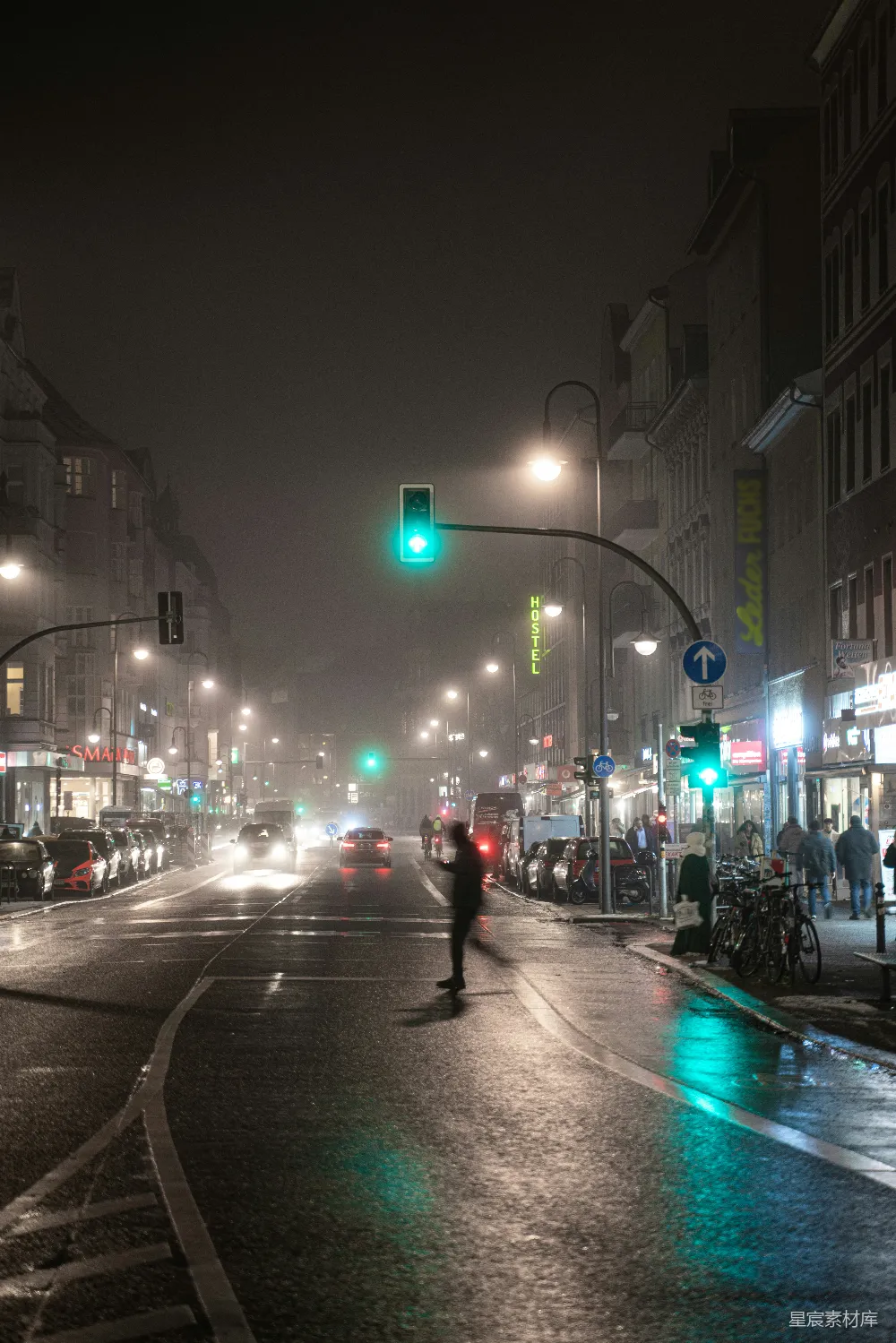 这张图片展示了一个雨夜中的城市街道场景。街道两旁的建筑物灯火通明，绿色的交通信号灯在夜色中显得格外醒目。地面上的积水反射着灯光，增添了一丝湿润与凉意。行人和车辆在这样的氛围中穿梭，构成了一幅忙碌而充满生活气息的夜景画面。