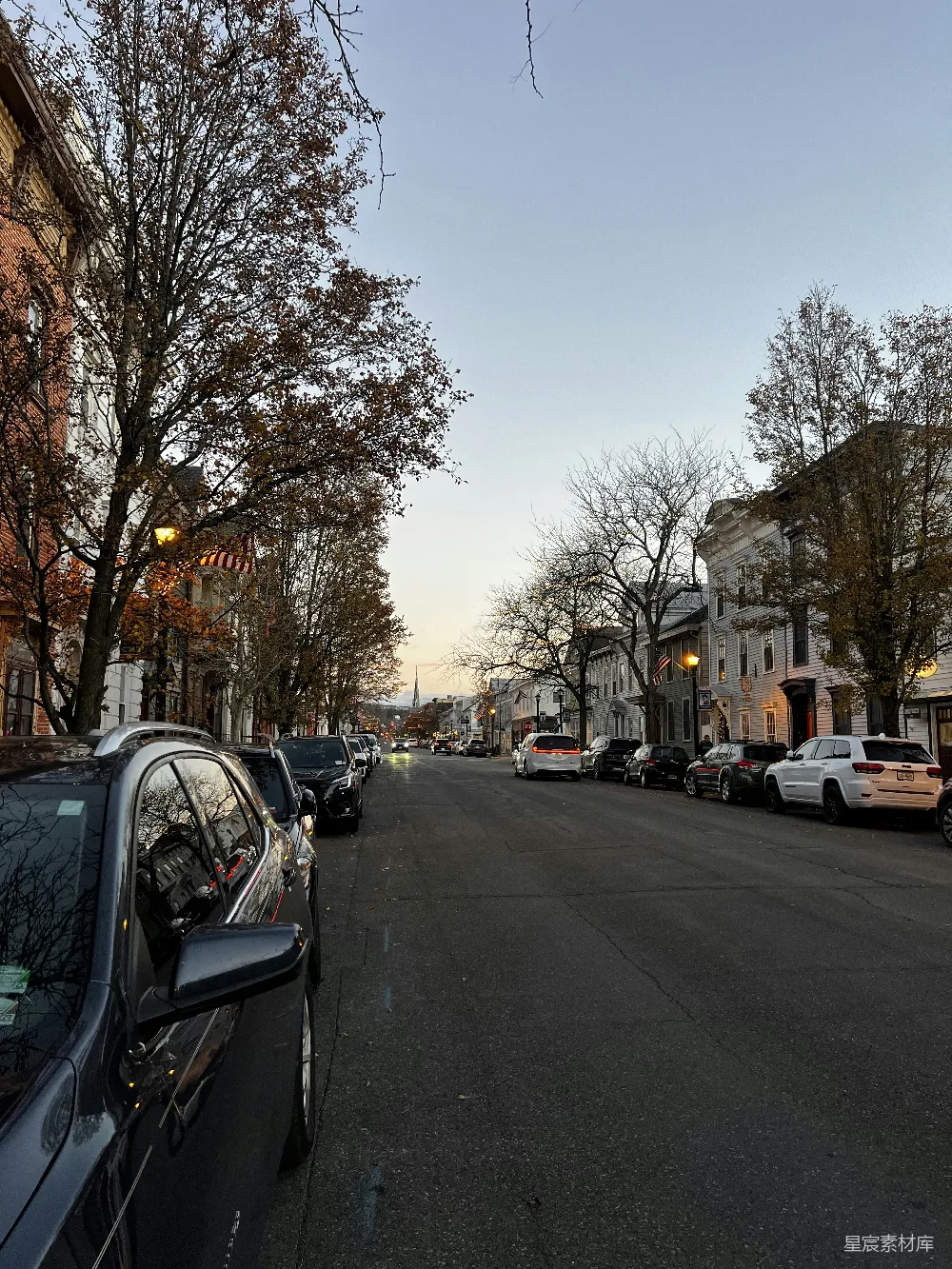 这张照片展示了一个黄昏时分的宁静街景。街道两旁是整齐排列的住宅楼，树木的枝叶在微风中轻轻摇曳，路灯的温暖光芒开始显现。街道上停放着几辆汽车，画面中没有行人，给人一种平和与静谧的感觉。适合用于展示城市生活的宁静时刻，或者作为城市街道的背景素材