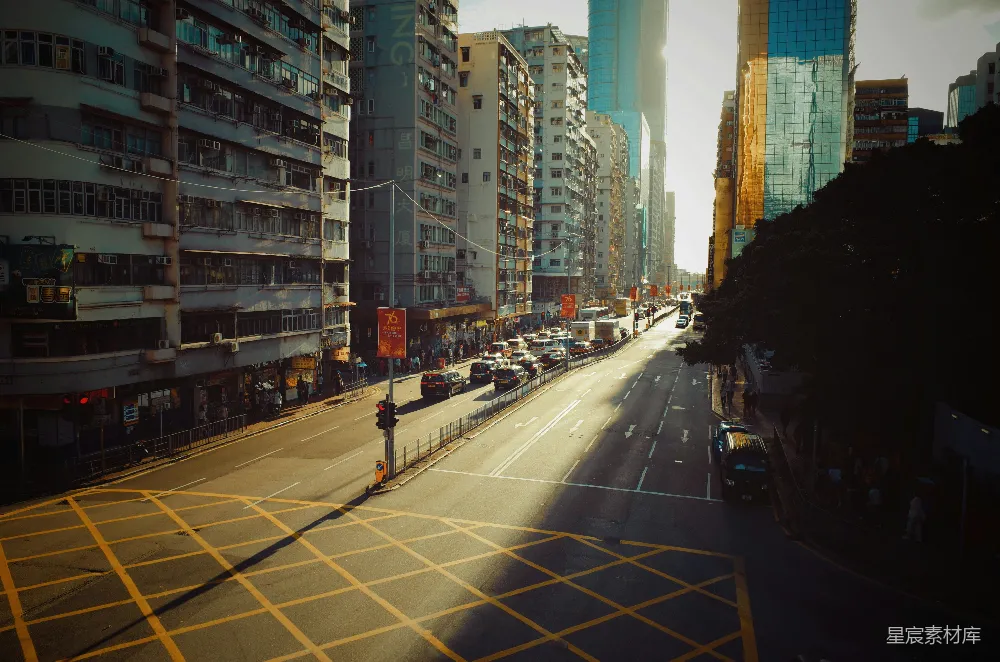 这张图片展示了香港的一条街道，夕阳的余晖洒在建筑物和路面上，形成了美丽的光影效果。两侧是现代化的高楼和老旧的居民楼，街道上车辆穿梭，行人稀少。整体画面给人一种宁静而充满生活气息的感觉。