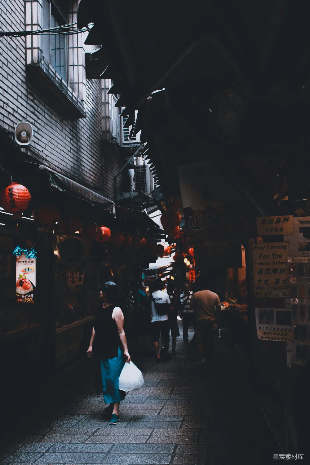 图片展示了夜晚昏黄的旧巷中，人们在市场中穿梭的场景。街道两旁是传统的商店，挂着红灯笼，营造出浓厚的市井氛围。画面色调偏暗，给人一种安静而温馨的感觉，适合用于展示城市生活或旅行相关的背景素材。
