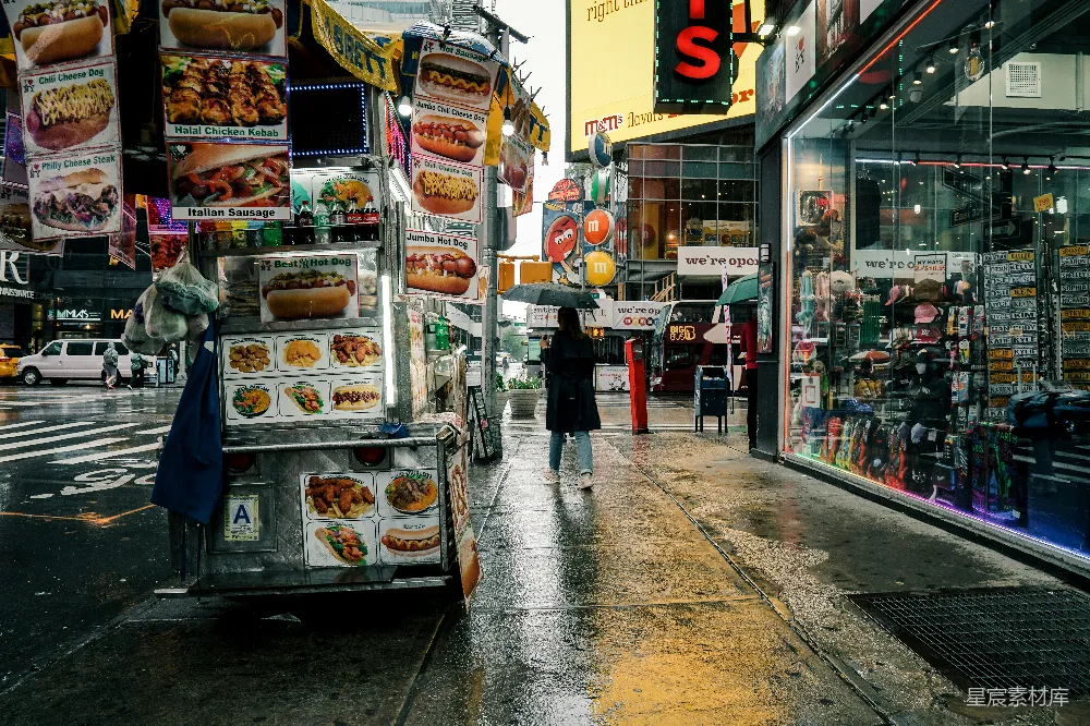 这张图片展示了一个雨中的纽约街头小吃车，摊位上挂着各种食物的招牌，吸引着路人的目光。街道两旁是现代化的建筑和商店，霓虹灯和广告牌在雨中显得格外醒目。一位撑伞的行人从小吃车旁走过，背景中可以看到行人和车辆，整个场景充满了都市生活的气息。