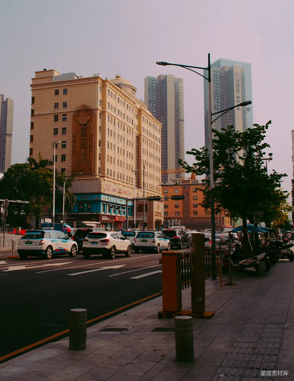 图片展示了一条繁忙的城市街道，背景是高耸入云的现代化高楼。街道两旁停满了车辆，路边设有橙色的交通隔离桩。远处的建筑风格多样，既有古典风格的建筑，也有现代化的高楼大厦，体现了城市的发展与变迁。