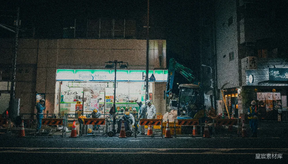 这张图片展示了一个夜晚的城市建筑工地场景。工人们在灯光照耀下忙碌地工作，背景中是一家FamilyMart便利店，周围堆满了施工材料和设备。整个画面给人一种忙碌而有序的感觉，展现了城市建设和生活中的一个侧面。