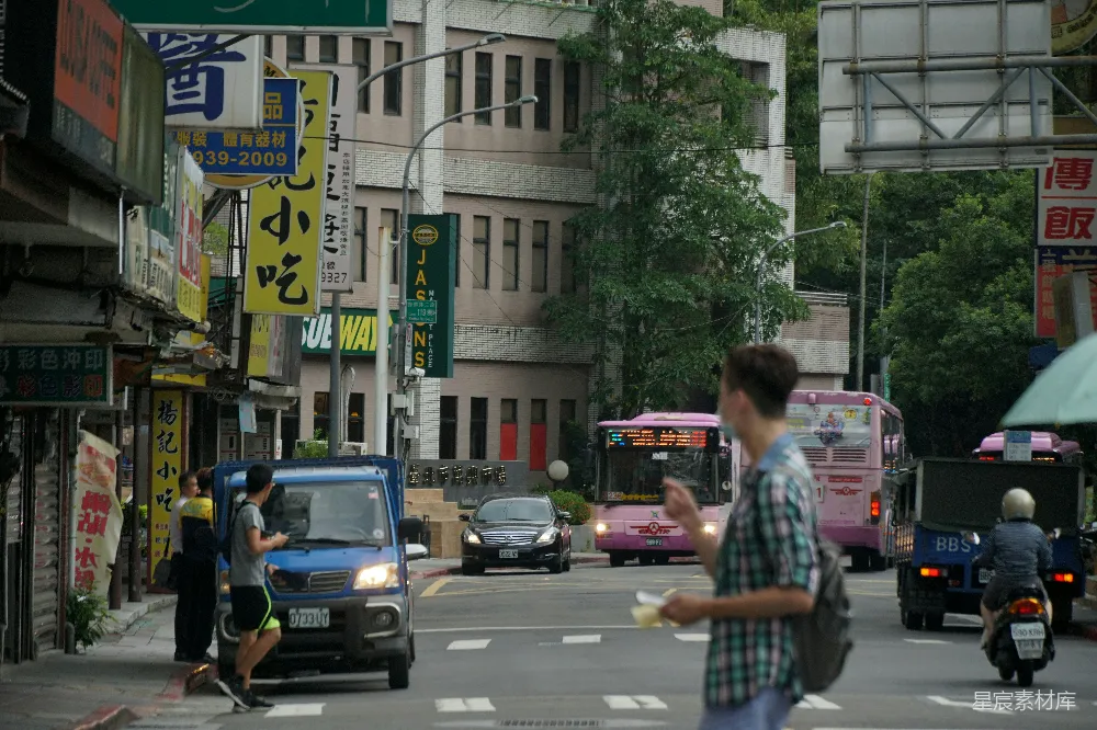 这张图片展示了一个繁忙的街道场景，车辆和行人穿梭其间，街道两旁是各种商店和广告牌，背景中有一栋建筑，树木郁郁葱葱。画面中的人们穿着休闲，有的在走路，有的在等待过马路，营造出一种忙碌而有序的城市生活氛围。