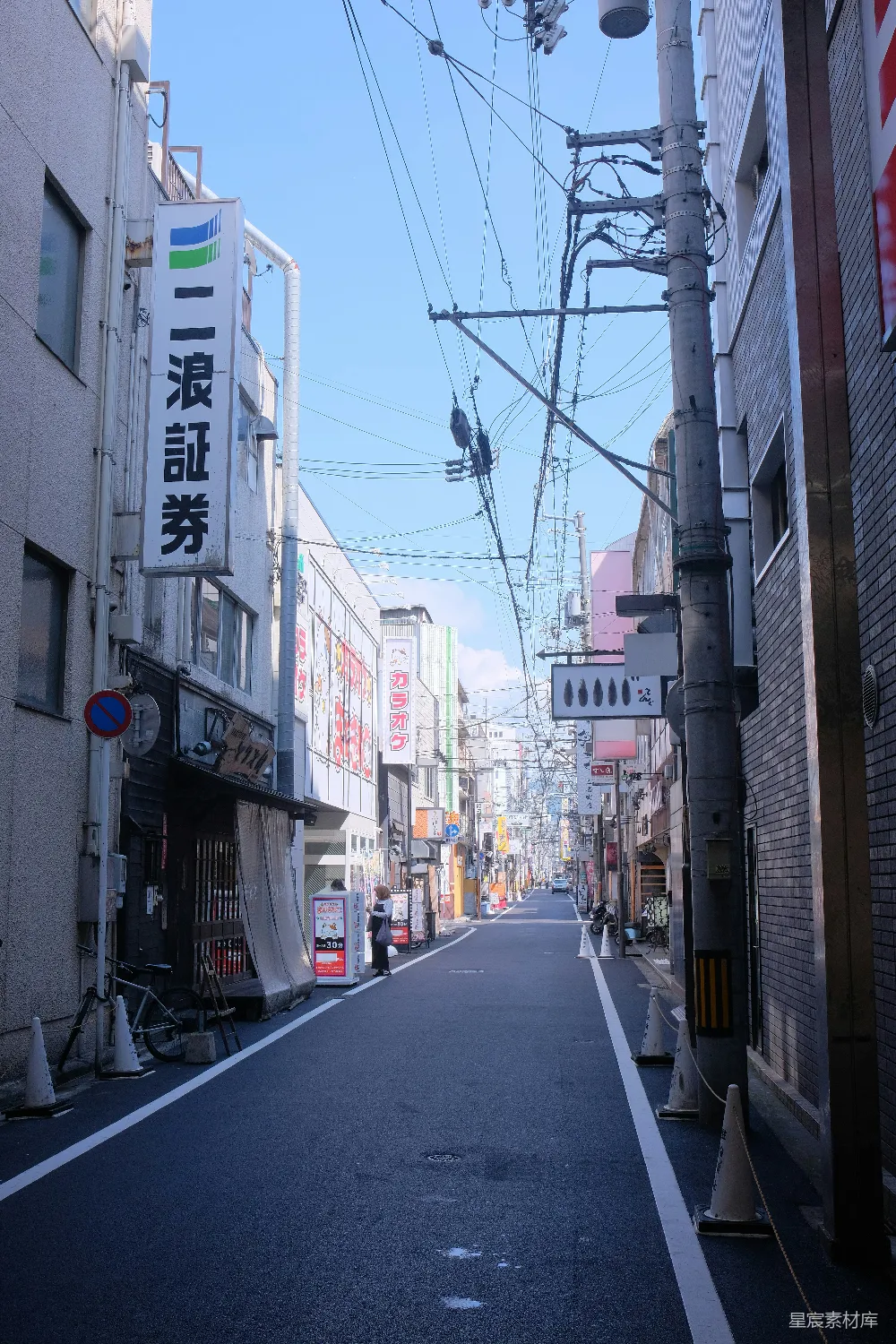 这张图片展示了一条狭窄的街道，晴朗的天空下，街道两旁是高大的建筑，电线杆上布满了各种电线。街道的尽头有一些商店，招牌上用日文书写。街道中间有一条自行车，地面上有交通锥，整体给人一种宁静而有序的城市生活印象。