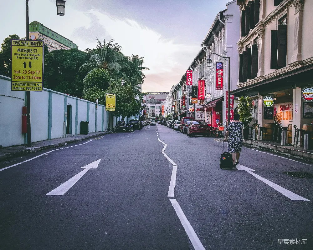 这张图片展示了一条安静的街道，两旁是风格独特的建筑，街道上空无一人，只有一个人拖着行李箱走在路上。街道两旁的建筑上挂满了红色的招牌，增添了浓厚的市井气息。天空呈现出美丽的渐变色彩，给人一种宁静而祥和的感觉。这张图片适合用于旅游宣传、城市风光