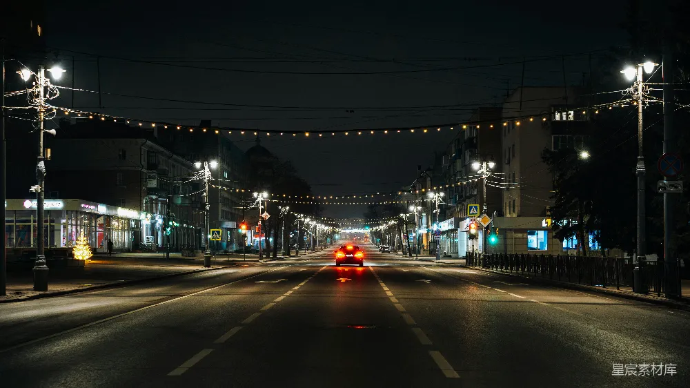 图片展示了一条夜晚的城市街道，街道两旁是高大的建筑物，街道上挂着一排排的灯饰，闪烁着温暖的光芒。远处有一辆红色的车正在行驶，车尾灯亮着，照亮了前方的道路。街道两侧的路灯和建筑物的灯光共同营造出一种宁静而神秘的氛围。这个场景适合用于描述城市夜