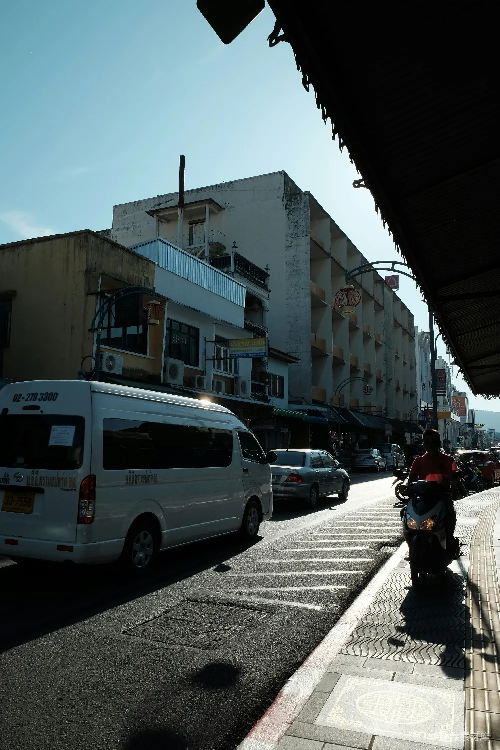 这张图片展示了一条城市街道的景象，阳光从建筑间洒落，照亮了行驶中的白色面包车和停在路边的摩托车。建筑物的外墙略显斑驳，显示出历史的痕迹。街道两旁停着几辆汽车，街道一侧有行人道，地面上有整齐的斑马线。整体画面给人一种宁静而繁忙的城市生活气息。