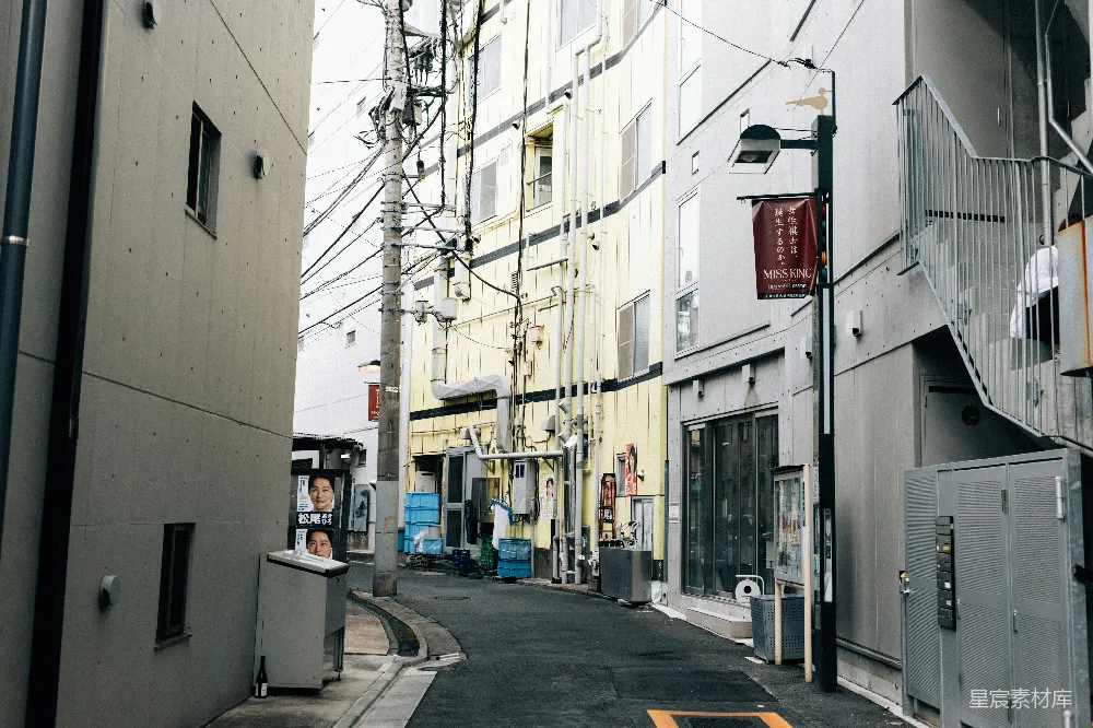 这张照片展示了一条典型的东京巷道，建筑物紧密相邻，电线杆上布满了各种电线，显得有些凌乱。巷道中有一些店铺和广告牌，墙壁上贴着一些宣传海报。整个场景给人一种繁忙而熟悉的都市生活氛围。