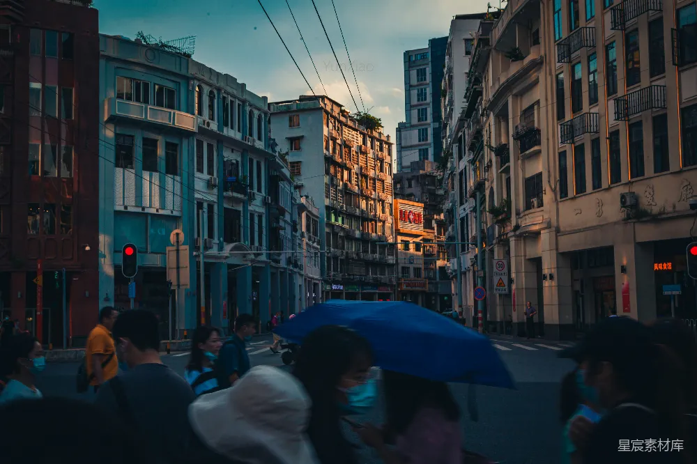 这张图片展示了雨中的城市街景，行人撑着伞走在路上，背景是不同风格的建筑。天空阴沉，街道上有一些行人戴着口罩，营造出一种宁静而略带忧郁的氛围。整体色调偏冷，给人一种清晨或傍晚的感觉。