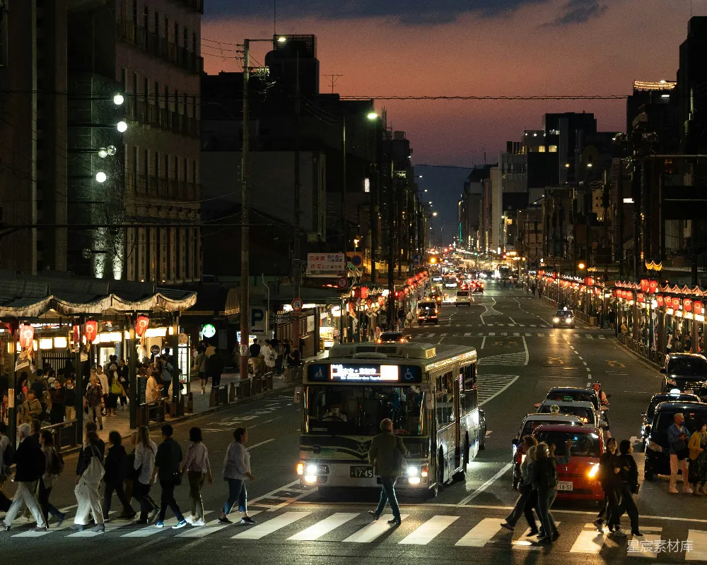 这张图片展示了夜晚繁忙的城市街道景象。街道两旁是高大的建筑物，灯光璀璨，行人和车辆络绎不绝。远处的天空被夕阳染成深红色，给人一种宁静而繁忙的感觉。这个场景非常适合用来表现城市生活的繁华与活力。