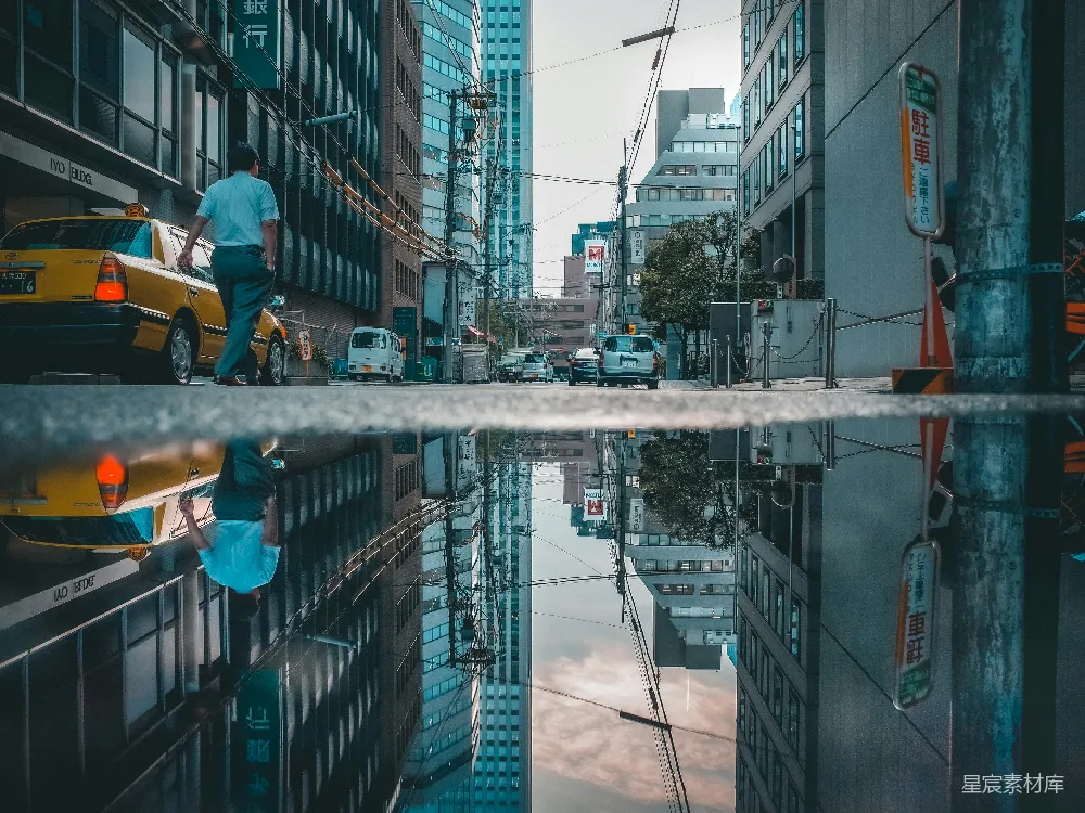 这张图片展示了城市街道的倒影，通过镜子的反射，将对称的建筑和车辆映照出来，形成一种独特的视觉效果。背景是现代化的建筑和行驶的车辆，给人一种繁忙都市的感觉。适合用于展示现代都市的繁忙与倒影的美学。