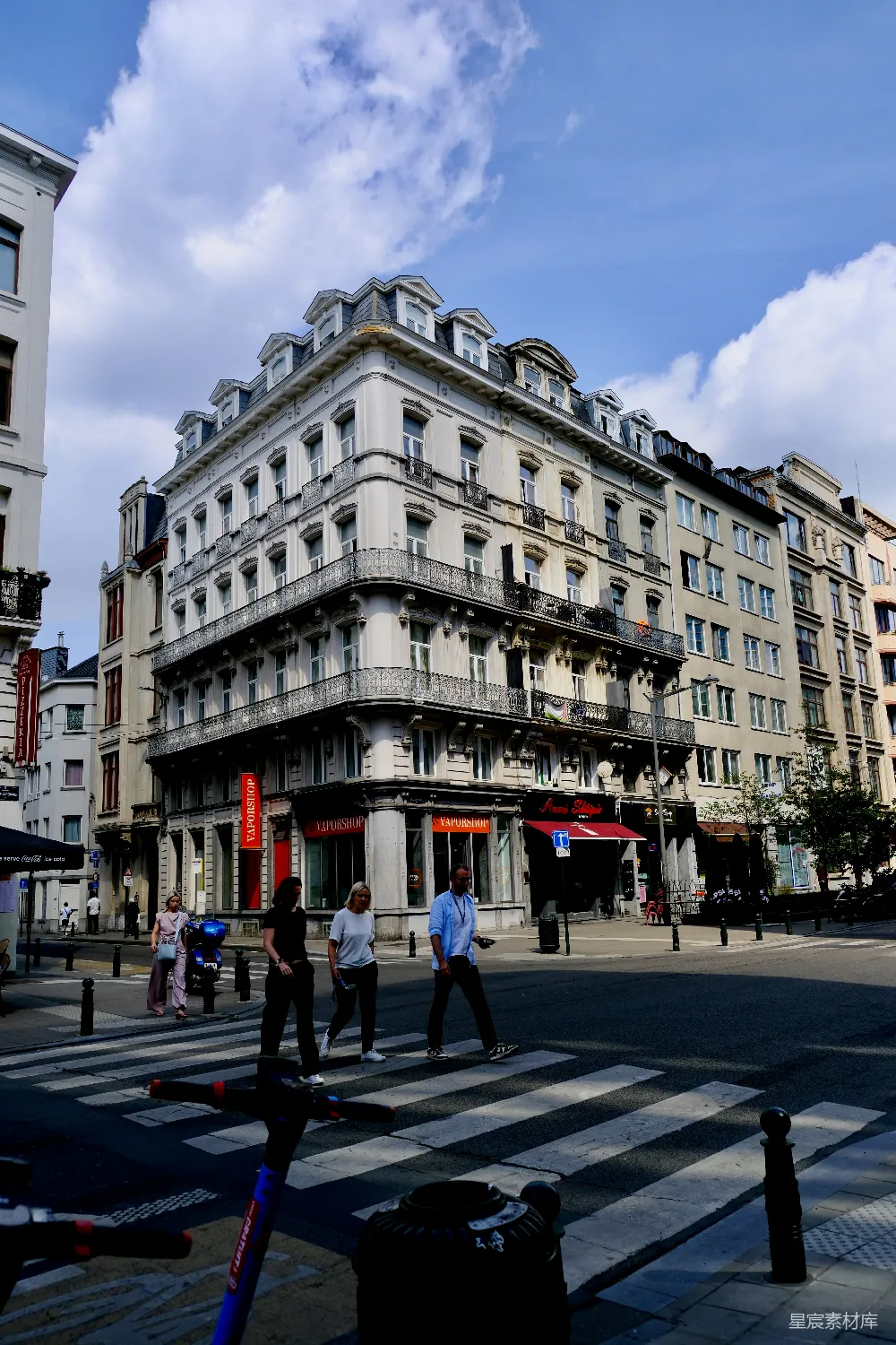 这张图片展示了一条欧洲城市的街道，背景是具有古典风格的白色建筑，带有精致的阳台和拱形窗户。前景中，现代的电动滑板车停在斑马线上，与行人和周围的环境形成了鲜明的对比。天空晴朗，云朵轻盈，增添了画面的生动感。这幅图像捕捉了城市生活中传统与现代的