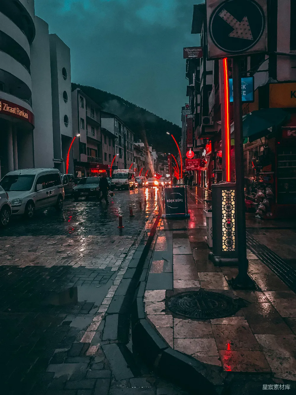 这张图片展示了一条充满霓虹灯的城市街道，在雨夜中显得格外迷人。街道两旁的建筑被灯光照亮，地面上的雨水反射出五彩斑斓的光芒，营造出一种独特的氛围。街道上偶尔有行人和车辆经过，远处的山峦隐约可见，给人一种宁静而神秘的感觉。