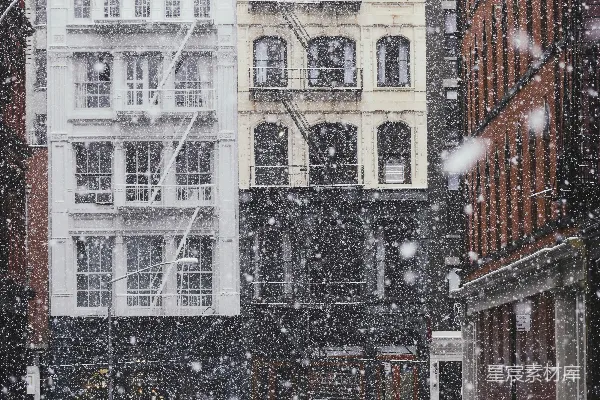 高清摄影 黑伞下的人站在覆盖着白雪的灰色沥青路上，靠近一幢商业建筑和一些汽车处，白天。