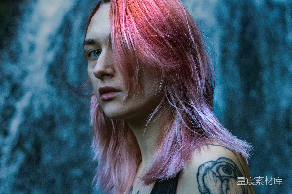 高清摄影 一头粉色头发并佩戴着耳饰的女人站在瀑布前。