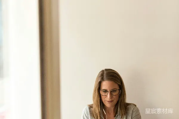 高清摄影 身穿灰色条纹衬衫的女士坐在棕色木桌前，旁边有一台白色的笔记本电脑，她正低头坐着。