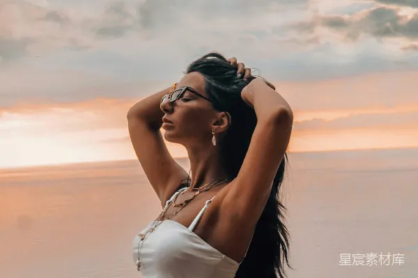 高清摄影 白T恤着装的女人站在海滩岸边，在日落时分穿着蓝色牛仔短裤站立。