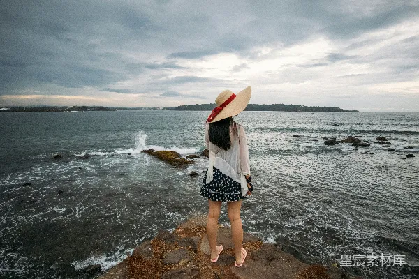 高清摄影 戴着帽子的女人在岩石海岸观看着海浪。
