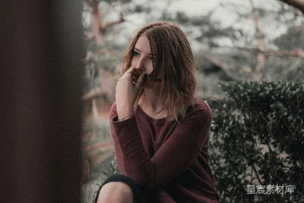 高清摄影 白天宽镜头摄影中，一位女性坐在 beige 色的木框架附近，背景是绿色灌木丛，天空有灰色的云朵。