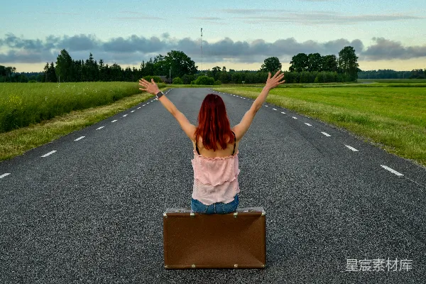 高清摄影 女人举起双臂坐在行李箱旁，处于开阔的道路上。