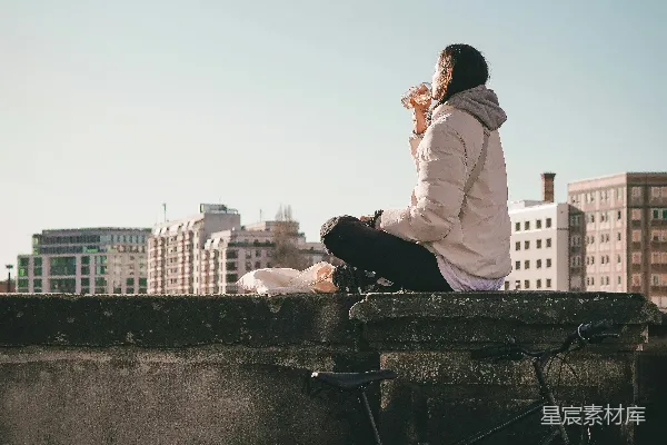 高清摄影 女人坐在墙角，脚踏自行车，背景是城市。