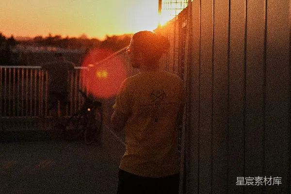 高清摄影 白圆领衫男穿黑裤站在栅栏旁，夕阳西下时。
