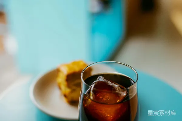 高清摄影 蓝桌上的玻璃杯中盛着一杯冰咖啡，旁边放着一块糕点。