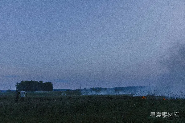 一个男子站在一片田野旁边，旁边有火光。