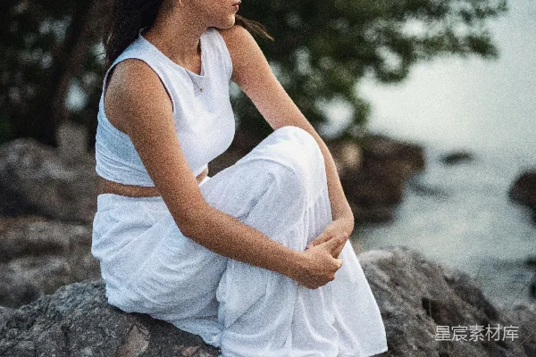 一个女人坐在一块石头上，旁边是一片水域。