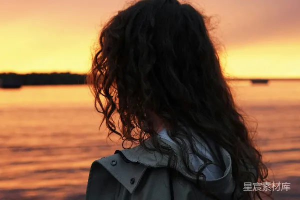 穿着灰大衣的女人站在海边，在夕阳西下时。
