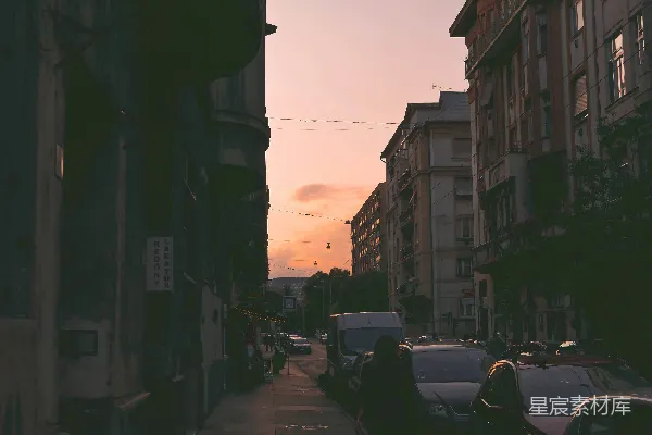 白天，在橙色天空下，人们在近建筑物的小道上行走，道路两旁有各种车辆。