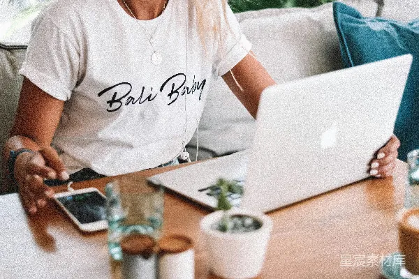 高清摄影 穿白色圆领T恤的女人坐在桌子旁，旁边有台MacBook电脑。